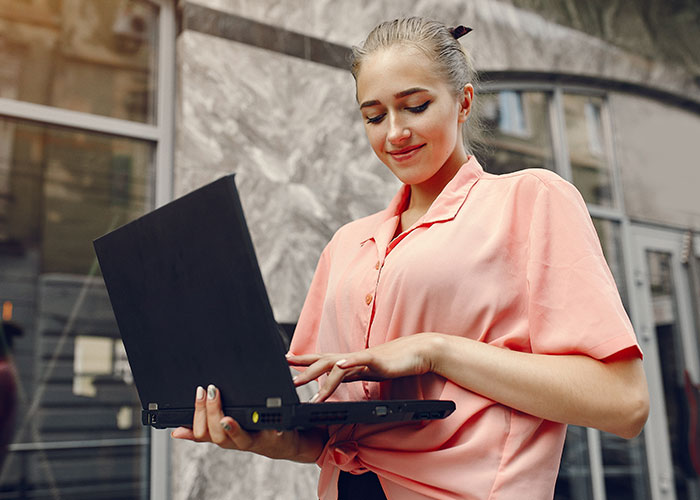 Woman in a pink shirt holds a laptop, smiling slightly, in a city setting; concept of mansplaining awareness.