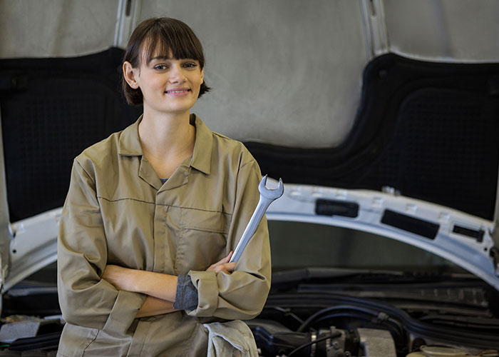 Woman in coveralls holding a wrench, standing by an open car hood, highlighting a mansplaining scenario in the garage.