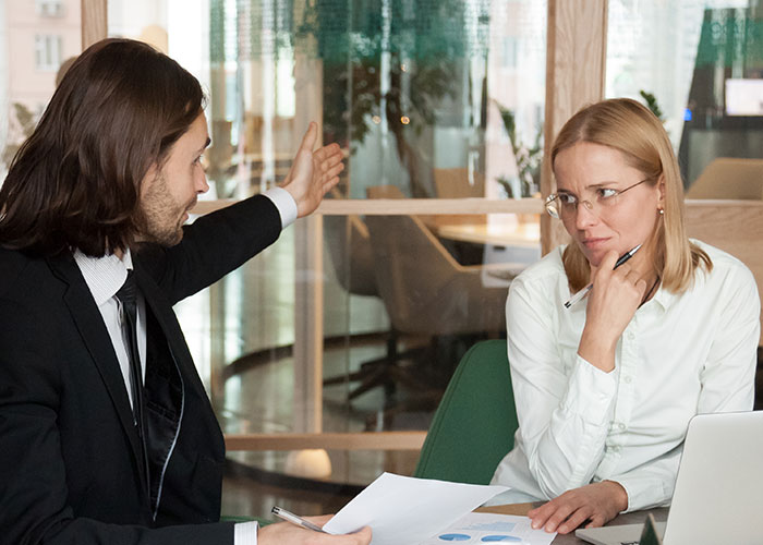 Man explaining at a desk, woman in glasses listening with skepticism, illustrating a mansplaining scenario in office setting.