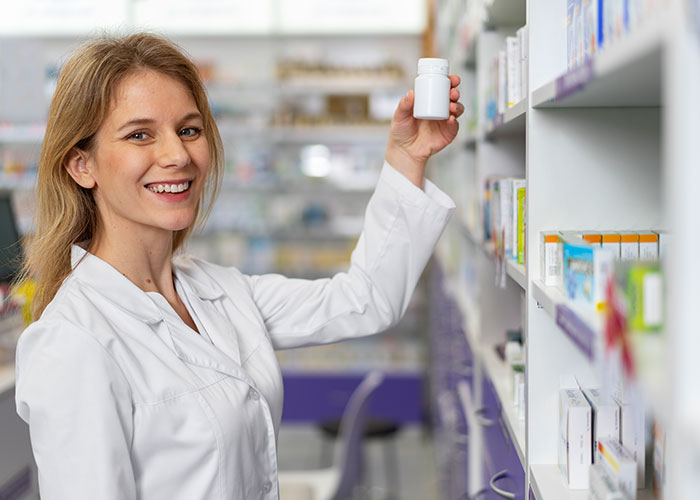 Pharmacist smiling while holding a white bottle in a pharmacy aisle, with shelves of medication visible.