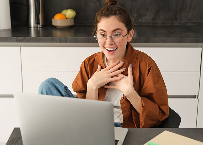 Woman in glasses and brown shirt looking amused at a laptop, reacting to mansplaining while in a modern kitchen.