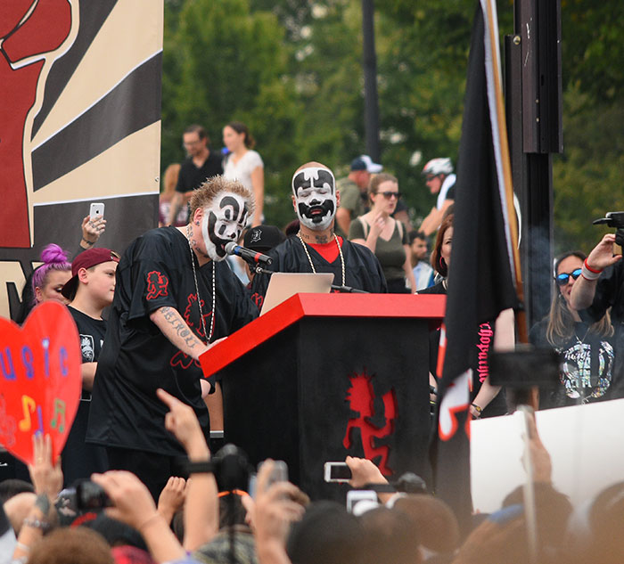 Two performers with face paint speak at a podium during an outdoor event, surrounded by a crowd.