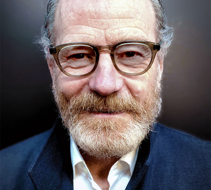 "Bearded celebrity with glasses and a navy jacket, smiling warmly at the camera."