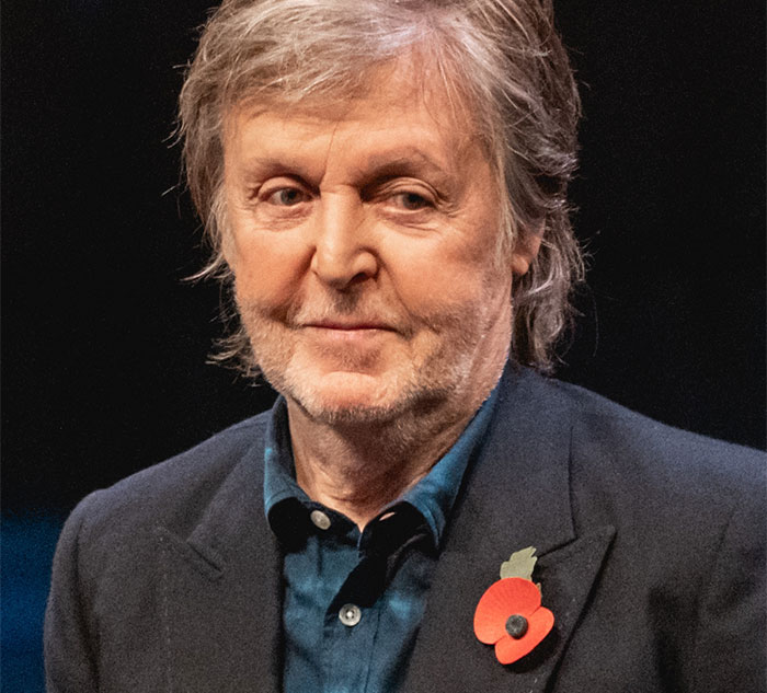 A celebrity in a suit with a poppy pin, smiling gently on stage.