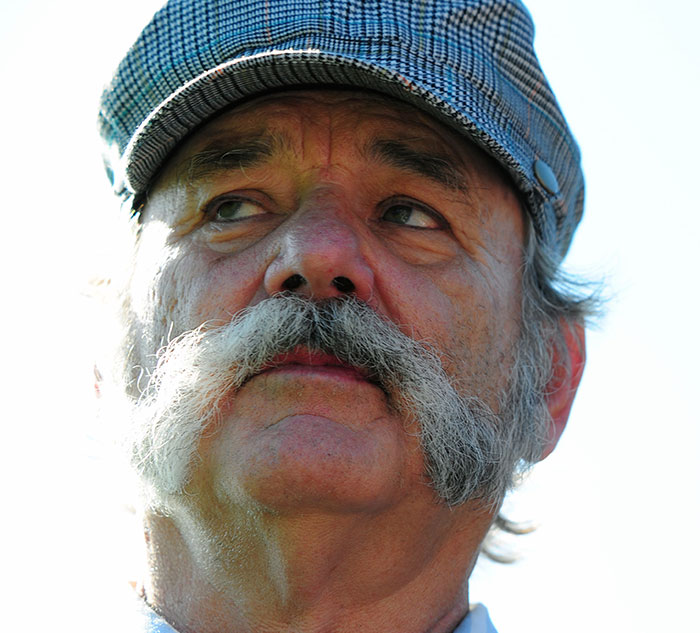 "Celebrity wearing a plaid hat, looking thoughtful with a bushy mustache, under a clear sky."