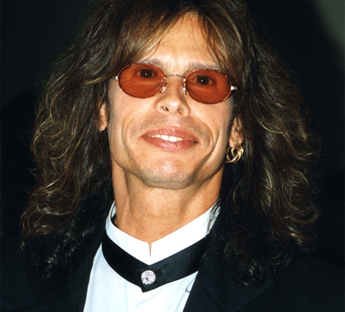 Celebrity in a tuxedo with long hair and orange-tinted glasses, smiling and looking at the camera.