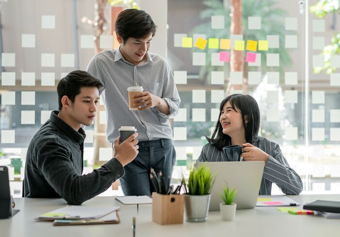 Three colleagues discussing job exits over coffee in an office with sticky notes on the window.