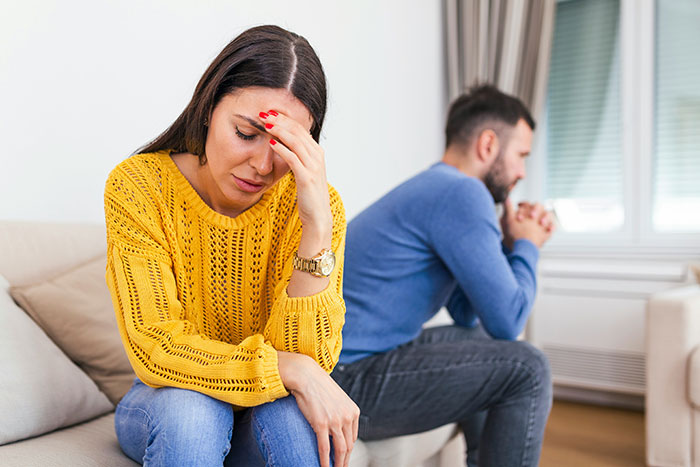 Woman in yellow sweater looking distressed, man in blue sweater sitting away, relationship conflict theme.