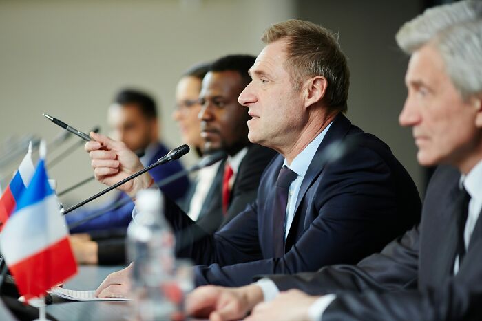 Businessmen in a panel discussion, focus on a man speaking, possibly addressing issues related to the biggest scam in history.