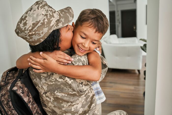 Soldier in uniform hugging smiling child in a living room setting.