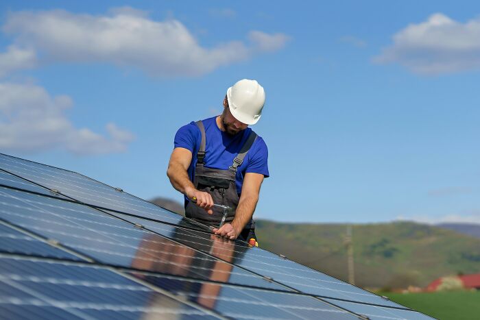 Worker in blue shirt and white helmet inspecting solar panels, blue sky in background.