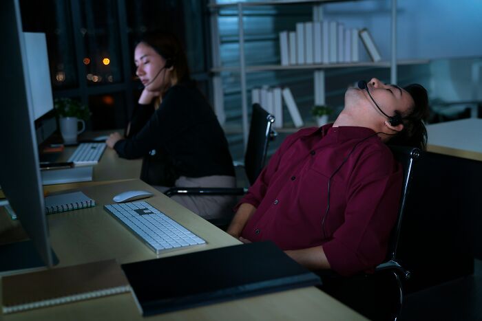 Two office workers at desks, one asleep, possibly involved in the biggest scam in human history, wearing headsets.