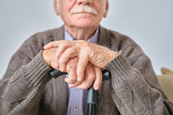 Elderly man sitting with a cane, hands resting on it, wearing a cardigan; symbolizing adulthood challenges.