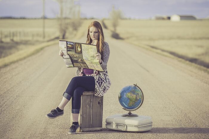 Woman with a map sitting on a rural road, suitcase and globe beside her, exploring travel choices.