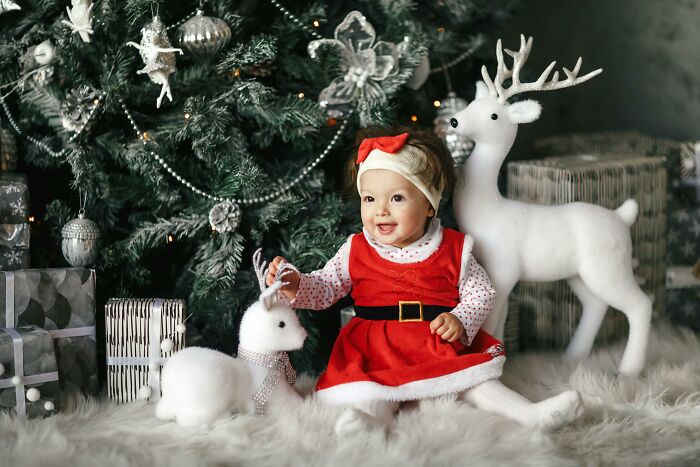 Baby in festive attire sitting by Christmas tree with gifts and reindeer, representing a babysitting offer from in-laws.