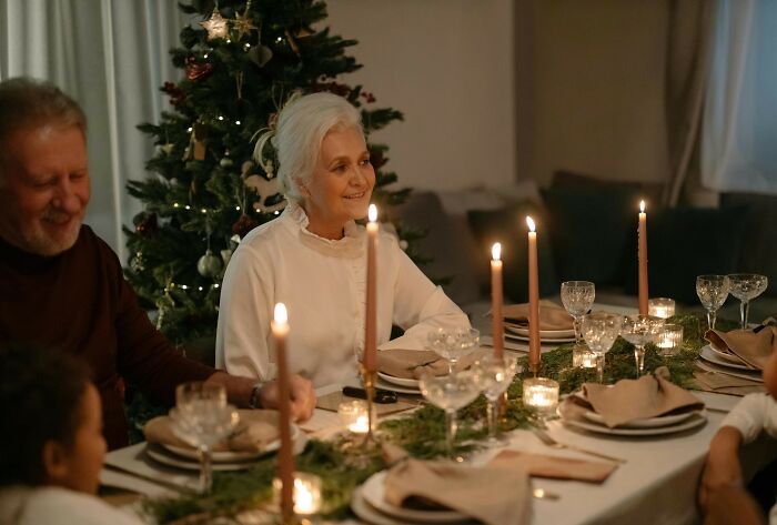 Elderly couple at a festive dinner table, with a Christmas tree in the background, discussing a babysitting offer.