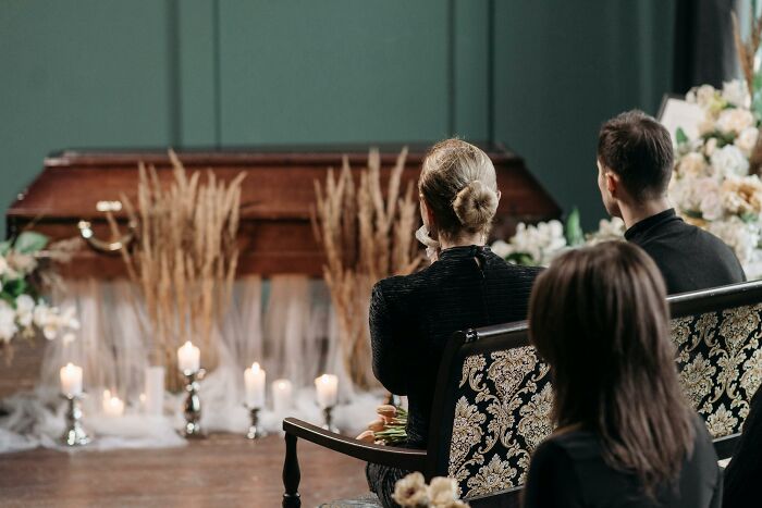 People in black outfits at a somber gathering, facing a casket surrounded by candlelit silverware.