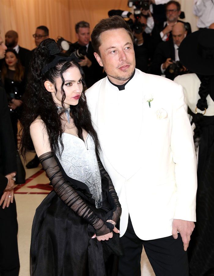 Couple posing at an event, with Grimes responding to controversial salute accusations; Elon Musk in a white tuxedo.