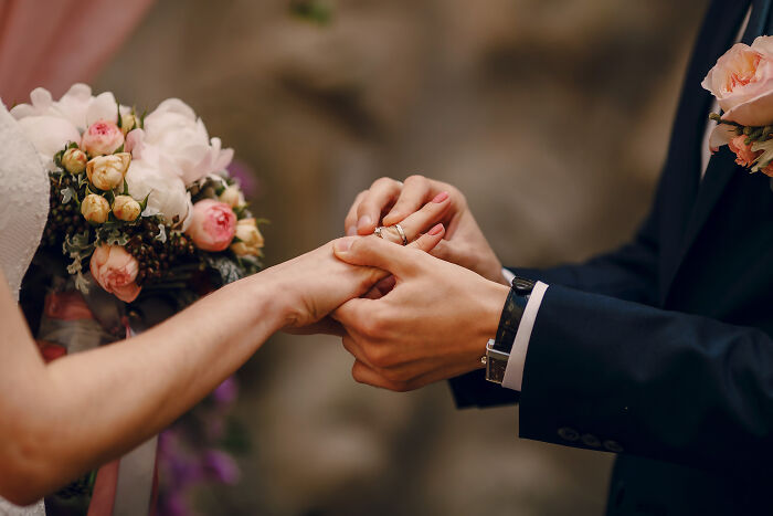 Couple exchanging wedding rings, highlighting unaffordable luxuries for the middle class with floral bouquet in background.