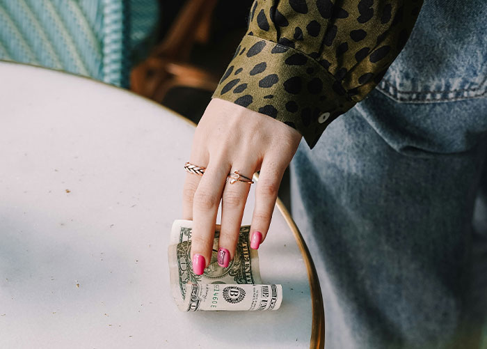 A person with pink nails holding a folded dollar bill on a table, suggesting wealth and ordinary moments.