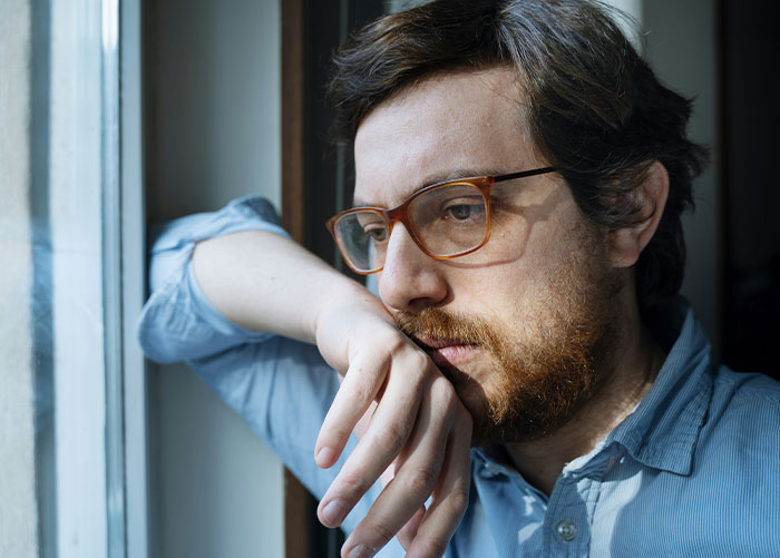 A thoughtful man with glasses looking out a window, reflecting on wealthy people's perceptions.