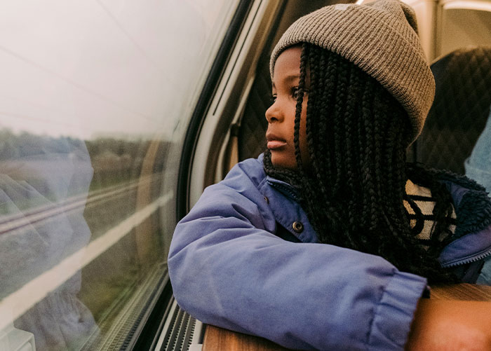 Child in a train, wearing a gray beanie and blue jacket, gazing out the window at the passing scenery. Wealthy lifestyle perspective.