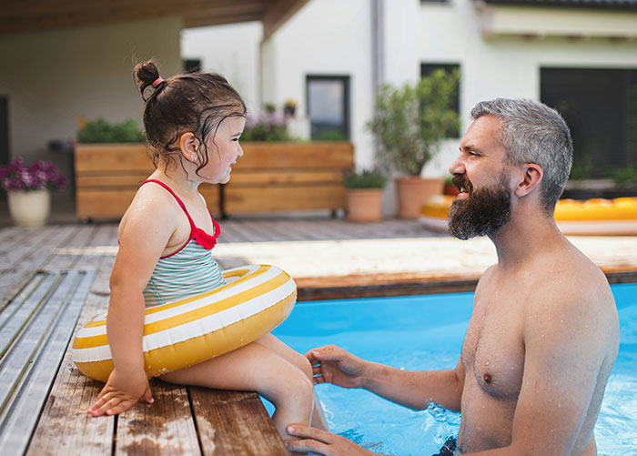 Man in a pool talks with a young girl sitting poolside in a swim ring.