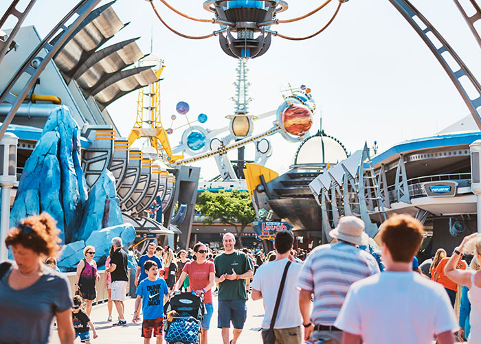 People strolling through a futuristic theme park on a sunny day, enjoying what appears to be ordinary attractions.