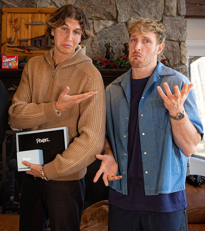 Two men posing humorously, one in a brown sweater, the other in a blue shirt, mimicking billionaire parodies.