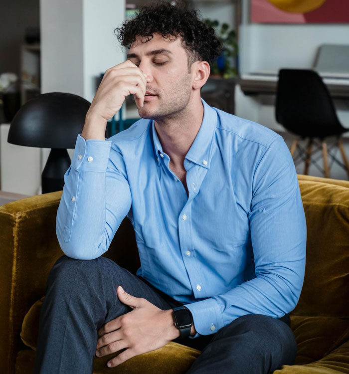 Man in a blue shirt sitting on a couch, appearing frustrated.