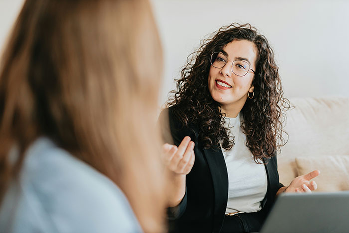 A woman with curly hair gestures while conversing about life lessons, sitting on a beige couch.