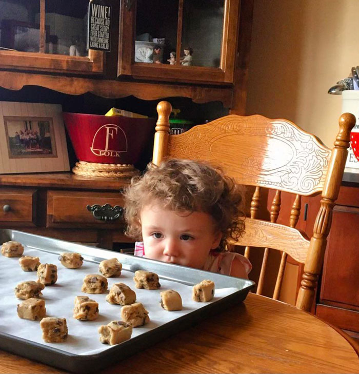 Curious child stares intently at a tray of cookie dough balls in a cozy kitchen setting. Hilarious kids moment.