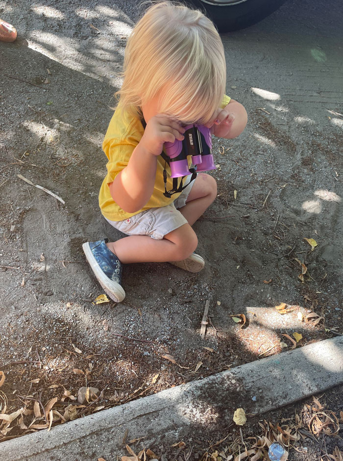 Child sitting on the ground wearing a yellow shirt and using purple binoculars, embodying a hilarious kids moment outdoors.