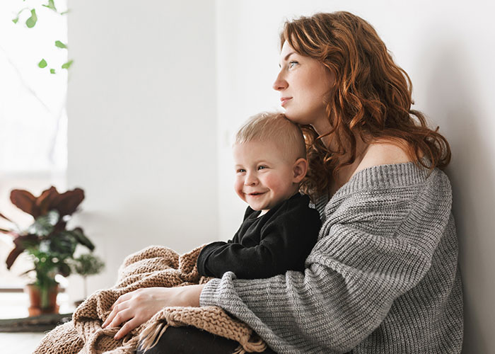 Woman holding a smiling child, wrapped in a brown knit blanket, sitting in a cozy room with plants.