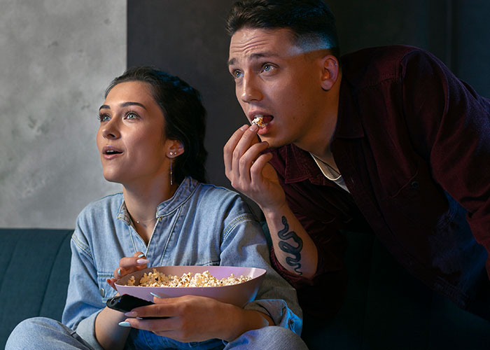 Couple eating popcorn, captivated by TV, unaware of the horrific reality behind normal things.