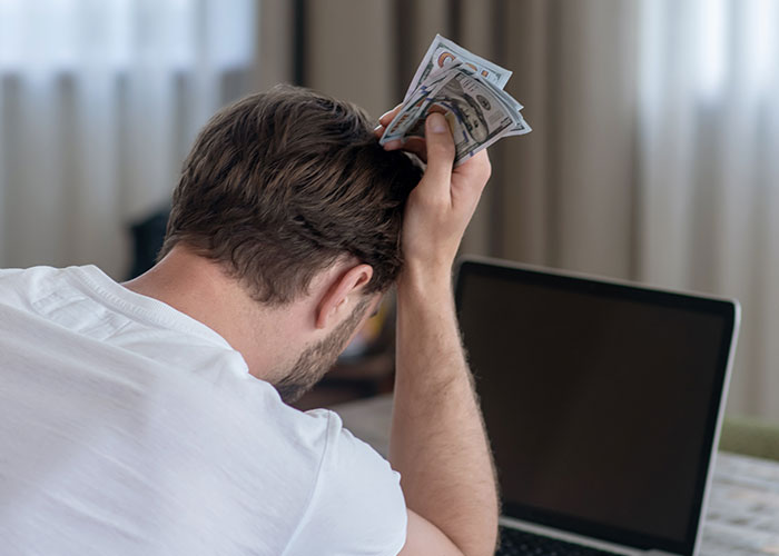 Man holding money in distress in front of laptop, highlighting the horrific side of normal financial stress.