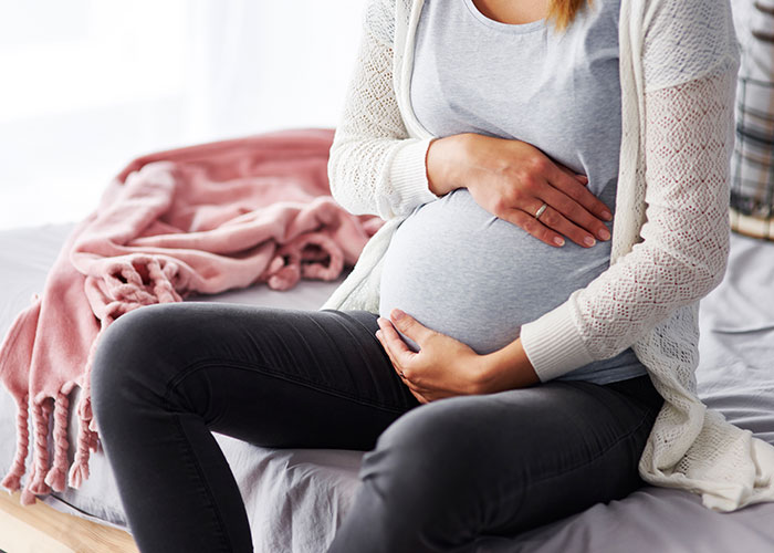 Pregnant woman sitting on a bed, cradling her belly, representing normal things that are deeply complex.