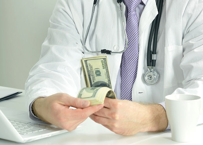 Doctor counting money at a desk with a stethoscope, reflecting on normal things that seem horrific.