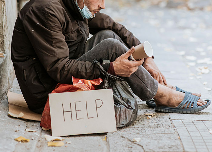 A person experiencing homelessness sits on the street with a sign reading "HELP" and a cup for donations.