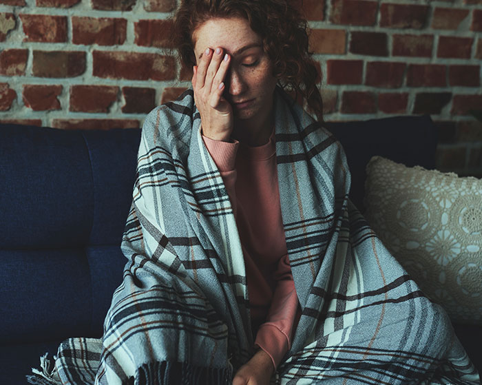 Woman wrapped in a blanket sitting on a couch, appearing thoughtful; highlights normal things with a potentially horrific angle.