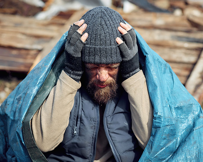 A person in worn clothing and gloves sitting under a tarp, illustrating a normal thing with horrific implications.