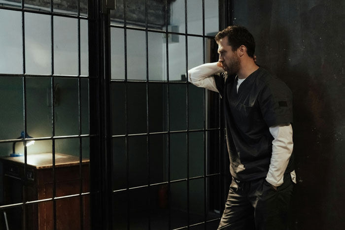 A man in scrubs standing pensively by a barred window, related to hospital expenses and childbirth.