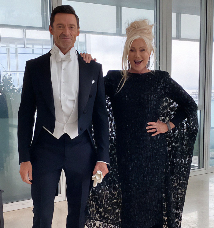 A man and woman dressed formally, posing indoors, with a backdrop of large windows.