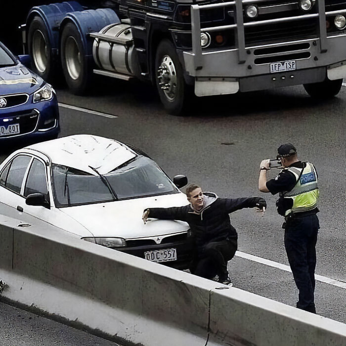 Man posing on freeway with a white car, while a police officer takes a photo; scene embodies humans-of-capitalism-pictures.