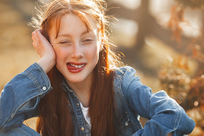 Girl in a denim jacket sitting outdoors, smiling playfully.