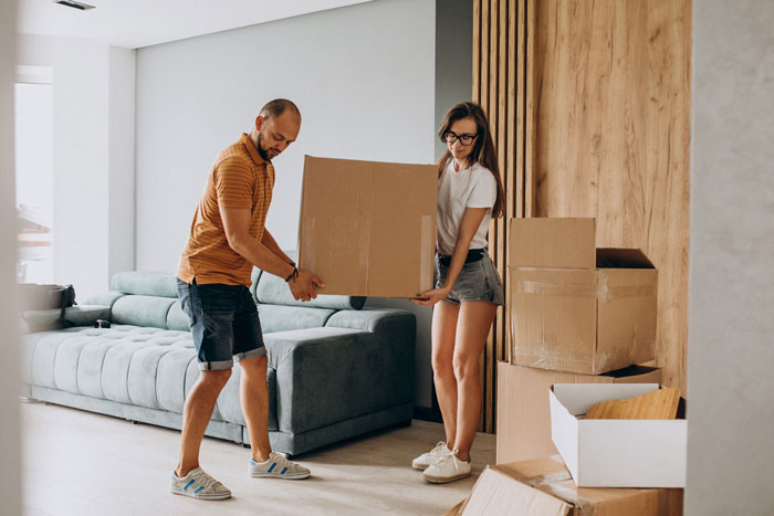 A man and woman lifting a cardboard box in a living room with packed boxes, considering legal action over unpaid bills.