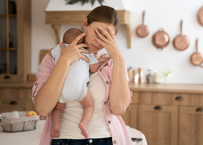 New mom looking overwhelmed, holding her baby in a kitchen with copper pans in the background.