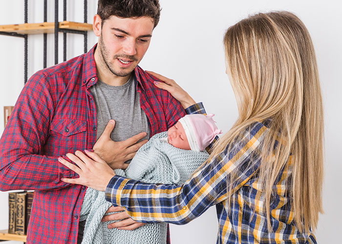 New mom looking overwhelmed, holding a baby, with husband in casual clothes.