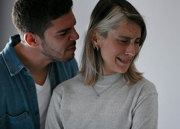 Overwhelmed new mom looks distressed as husband speaks close to her, both wearing casual clothes.