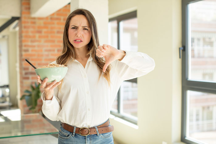 Woman expressing dissatisfaction in kitchen, holding bowl with thumb down, representing cooking drama.